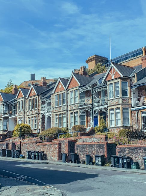 A row of Victorian-style terraced houses with multiple stories, decorative ironwork balconies, and bay windows, situated on a sloped street with a brick retaining wall and small landscaped gardens in 